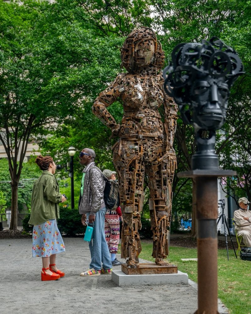 Close-up view of Olu Amoda's monumental scrap metal female sculpture, with visitors observing the artwork, and a black metal sculpture visible in the background
