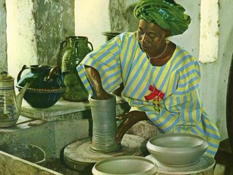 adi Kwali using a potter's wheel, wearing a striped blue and yellow traditional outfit with a green headwrap, surrounded by finished pottery pieces